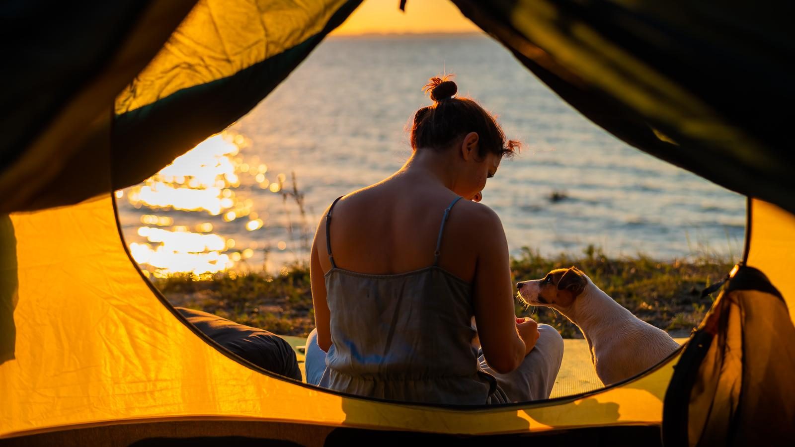Mujer con su perro sentada a la entrada de una tienda de campaña frente al mar al atardecer