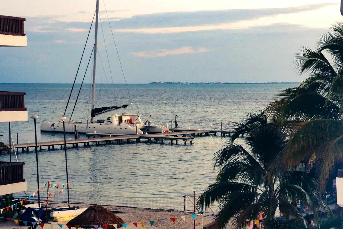Velero amarrado a un muelle en una bahía tranquila, visto desde un hotel con palmeras