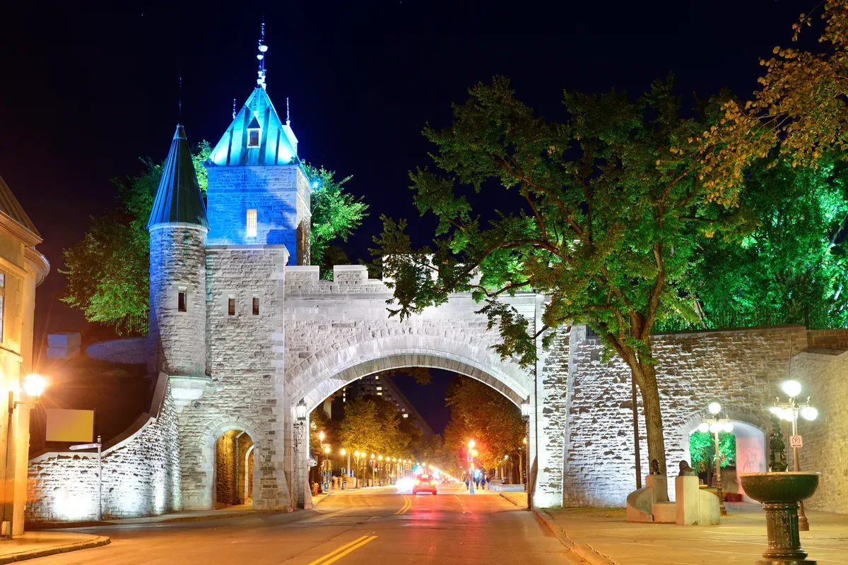 Puerta de piedra de una muralla con torreones iluminada de noche