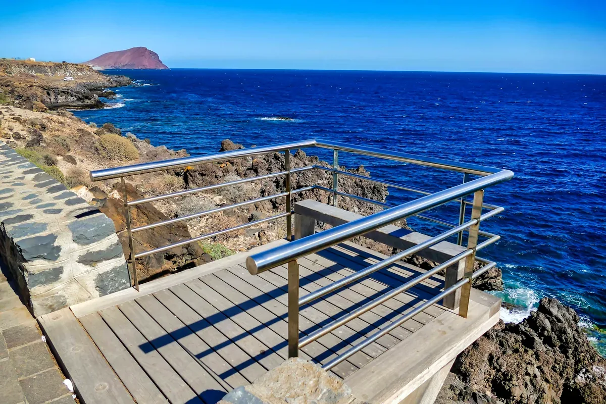 Mirador de madera con barandal frente a un mar azul y una costa rocosa