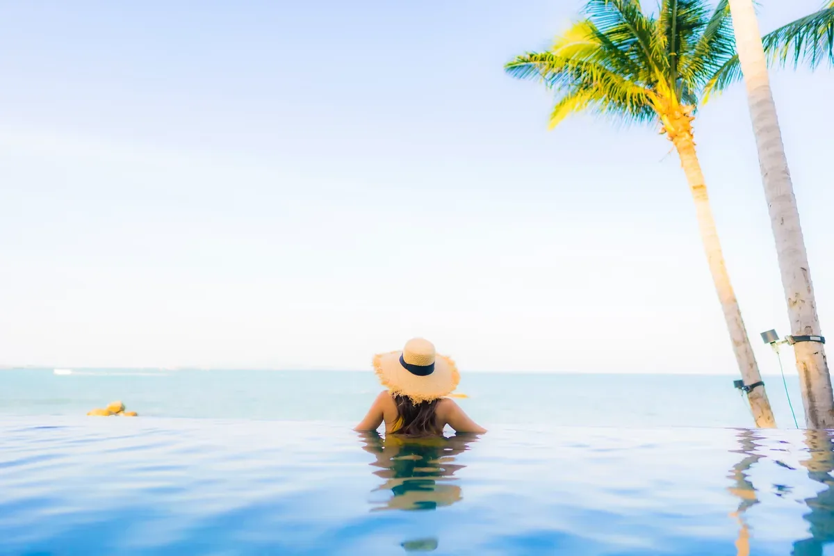 Mujer con sombrero de paja en una alberca infinita con palmeras y el mar al fondo