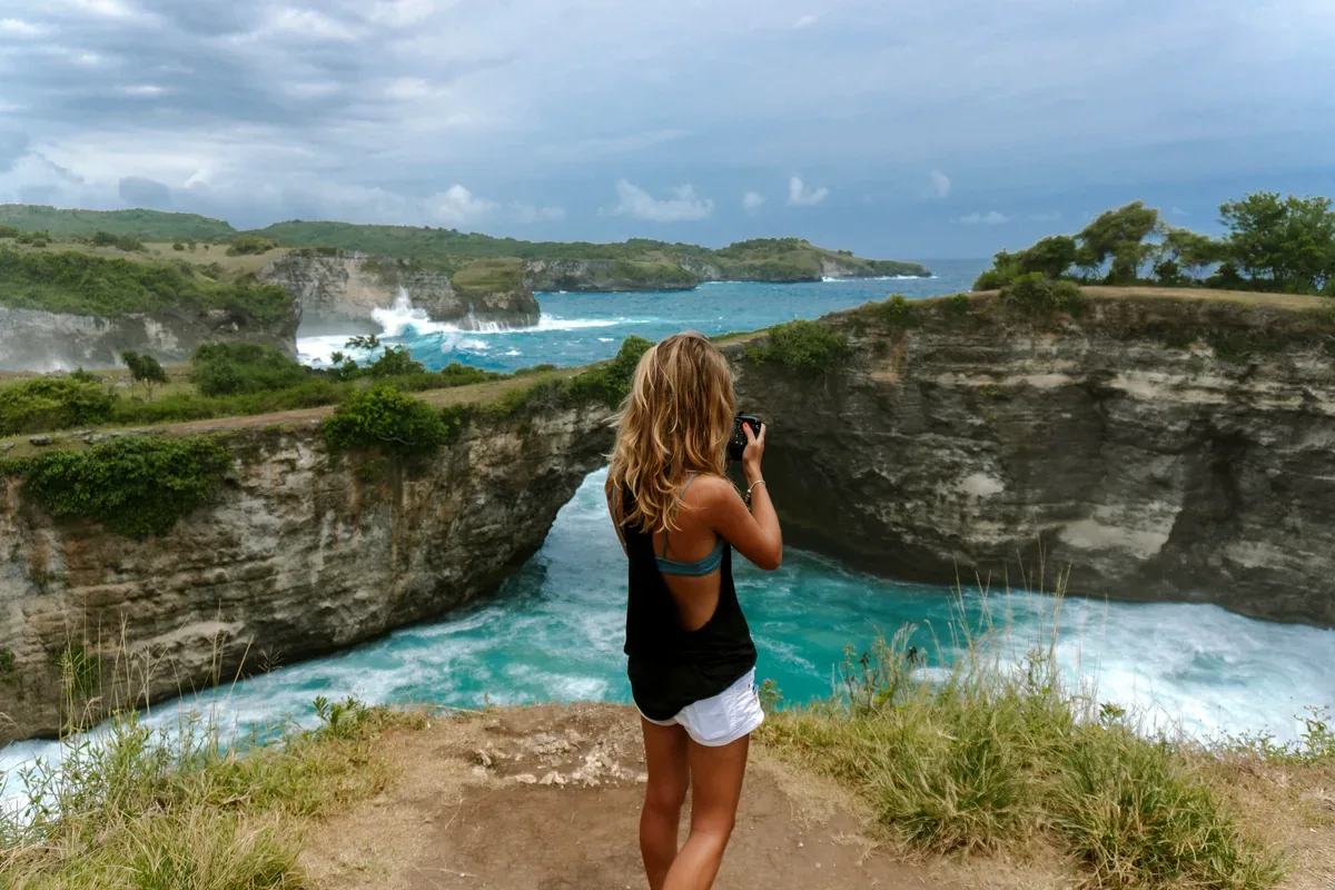 Mujer que toma fotos desde un acantilado frente a un arco natural de roca y el mar