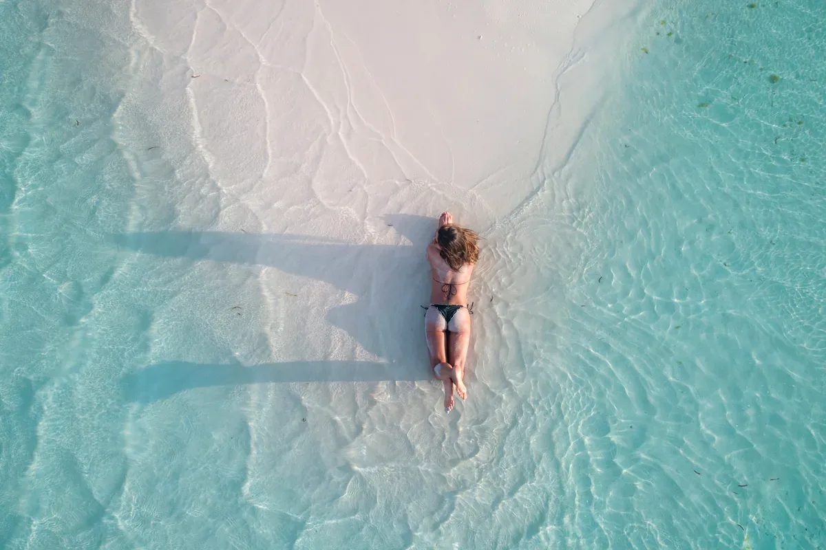 Mujer recostada en el agua transparente de una playa de arena blanca, vista desde el aire