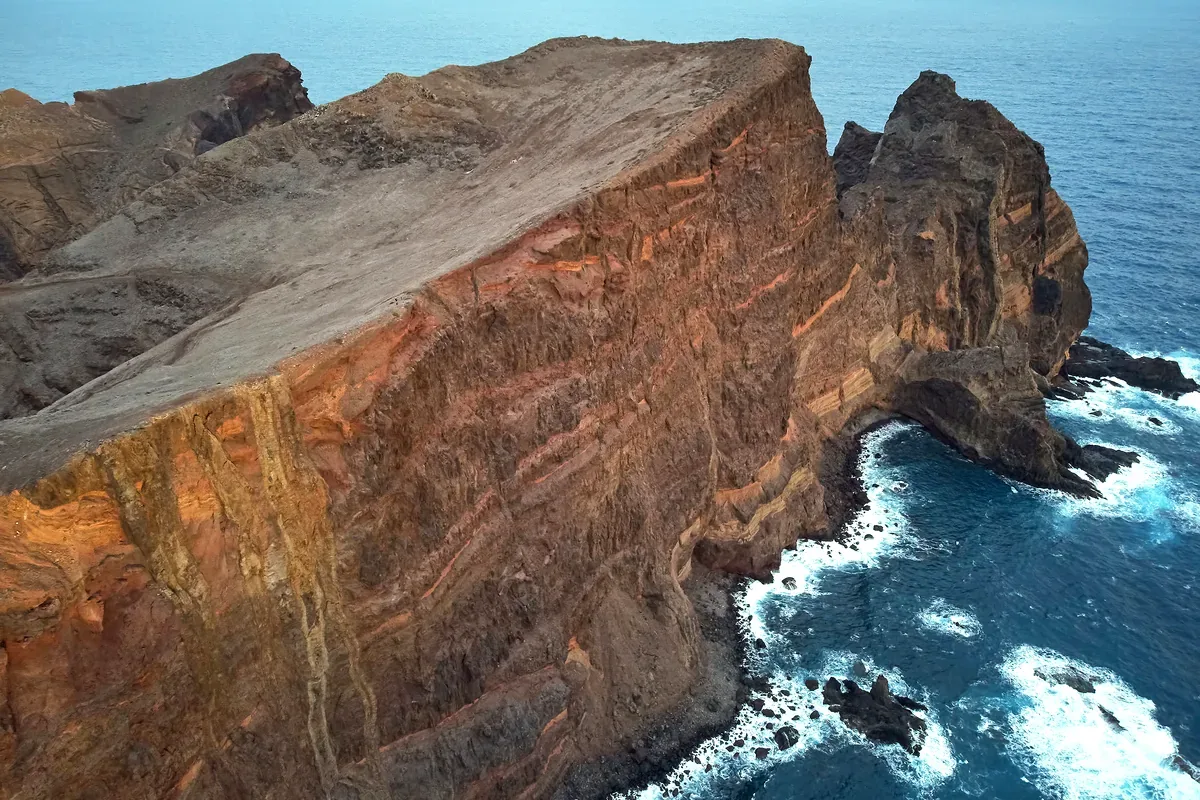 Acantilado de roca rojiza sobre el mar con olas que rompen en las rocas