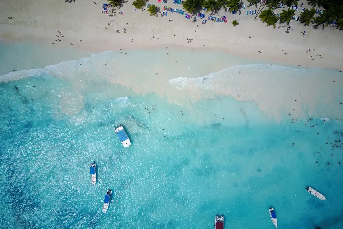 Lanchas en un mar turquesa frente a una playa con bañistas, vista desde el aire