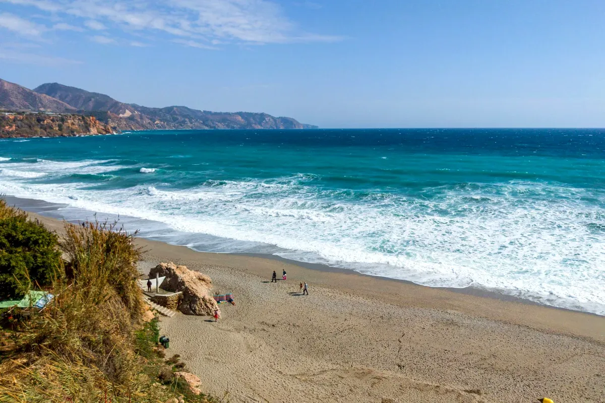 Playa de arena con olas y cerros costeros bajo un cielo azul