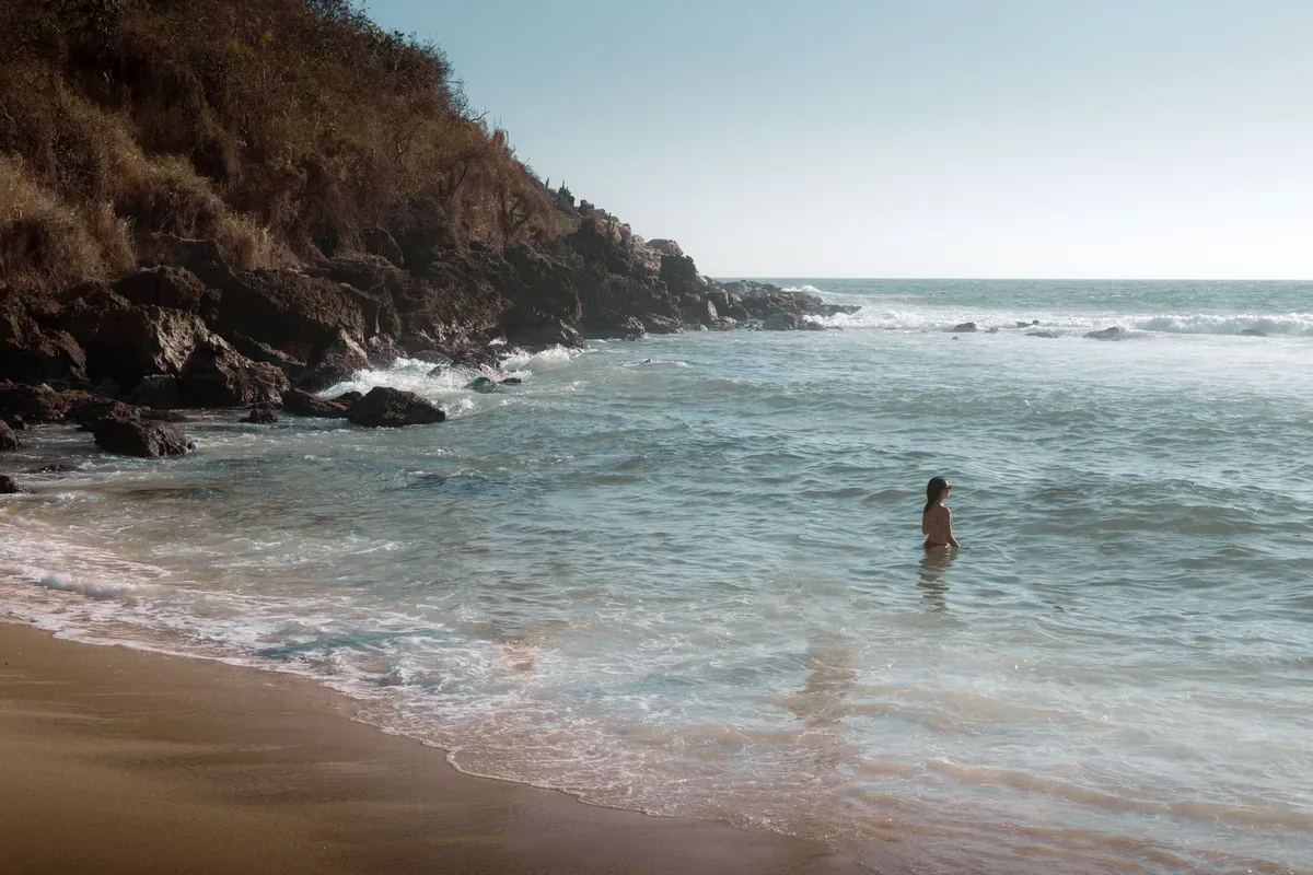 Persona dentro del mar en una playa de arena oscura al pie de un cerro rocoso