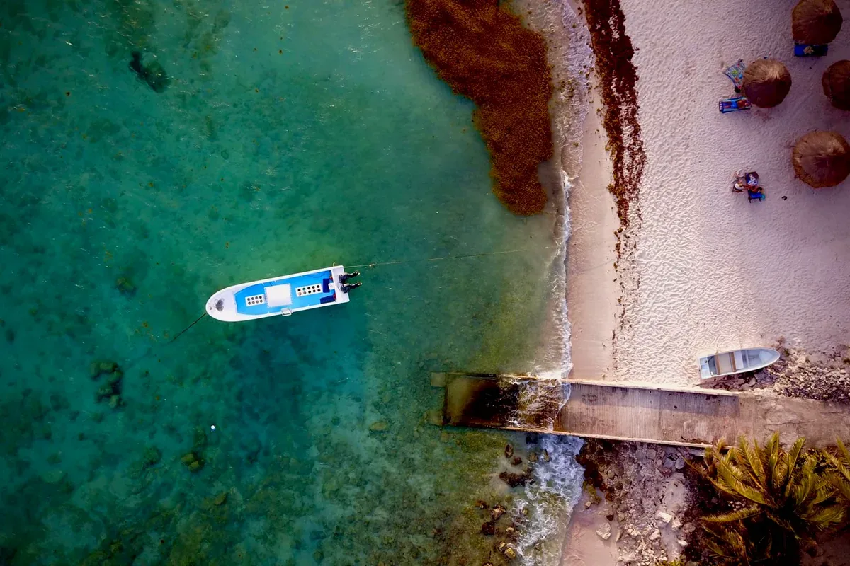 Lancha blanca fondeada en agua verde junto a un muelle y una playa