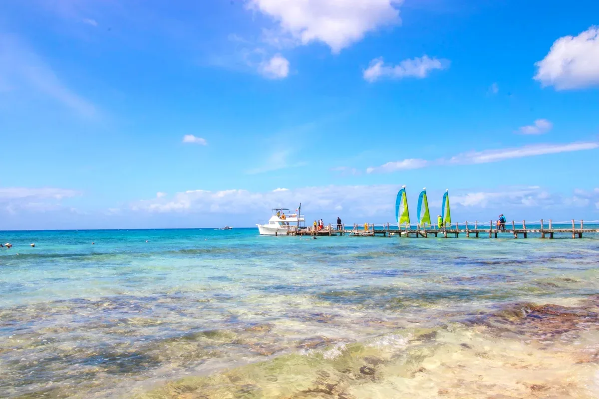Mar transparente con un muelle, lanchas y veleros de colores al fondo