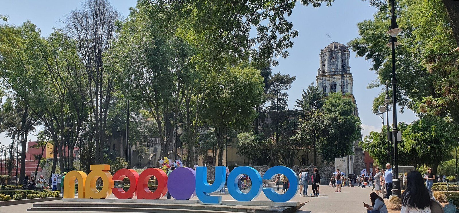 Letras de colores con el nombre Coyoacán en la Plaza Hidalgo, entre árboles, con la torre de la parroquia al fondo