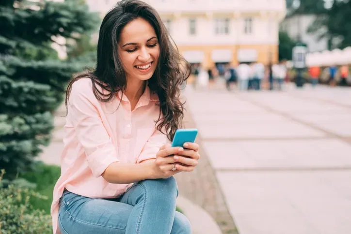 Mujer sonriente sentada en una plaza que revisa opciones de viaje en su celular