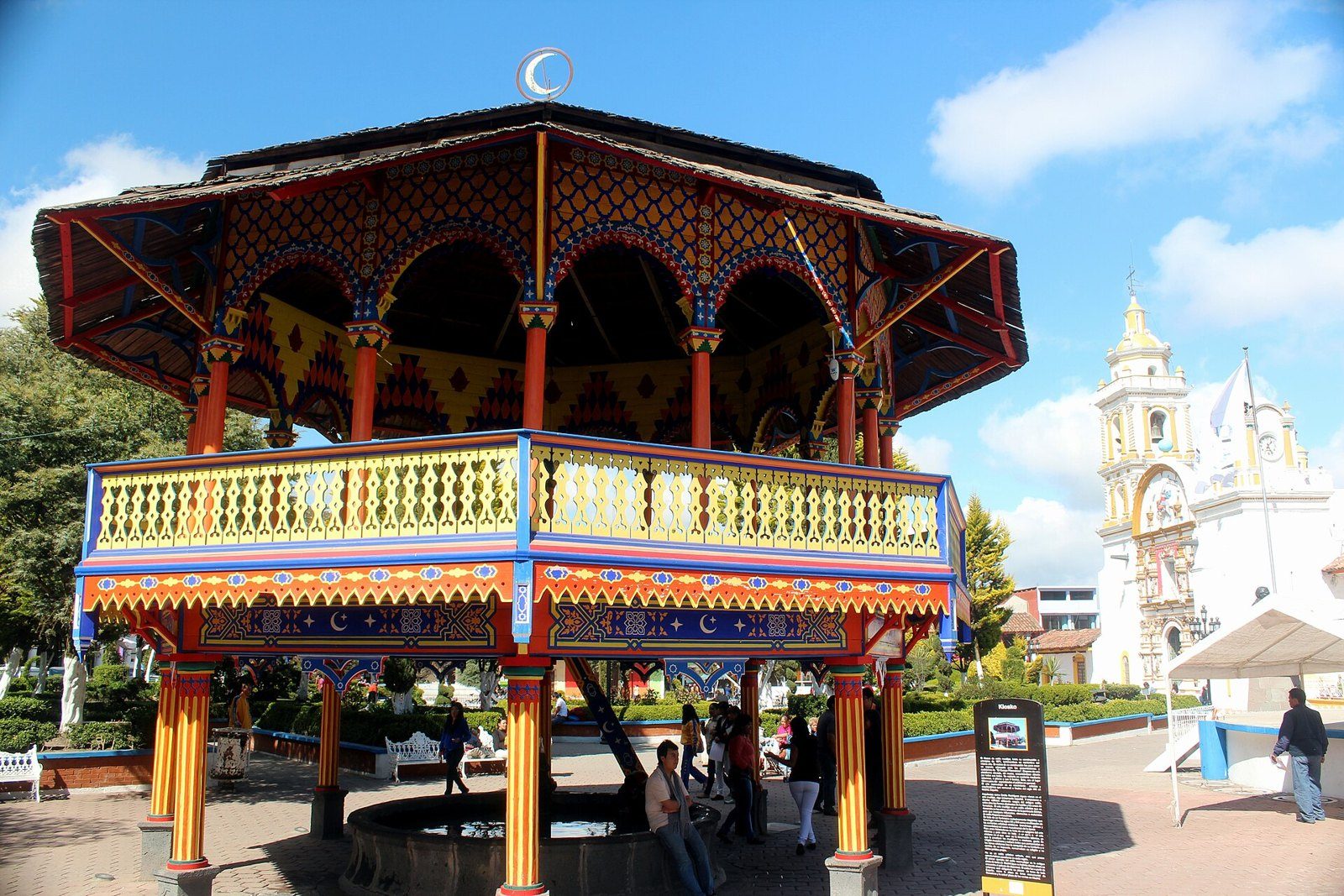 Kiosco de madera con barandal pintado de colores en la plaza de armas de Chignahuapan, con una iglesia de torre blanca al fondo