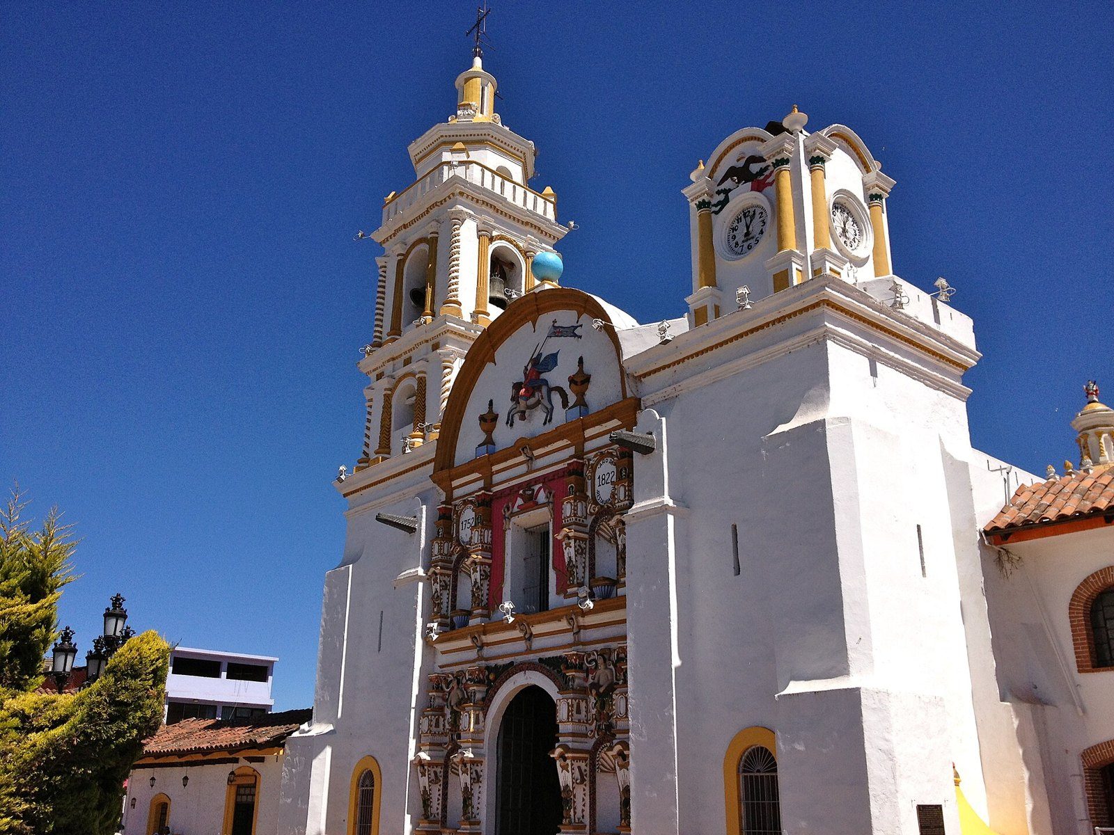 Fachada blanca de la Parroquia de Santiago Apóstol en Chignahuapan, con torre amarilla, reloj y portada decorada, bajo un cielo azul