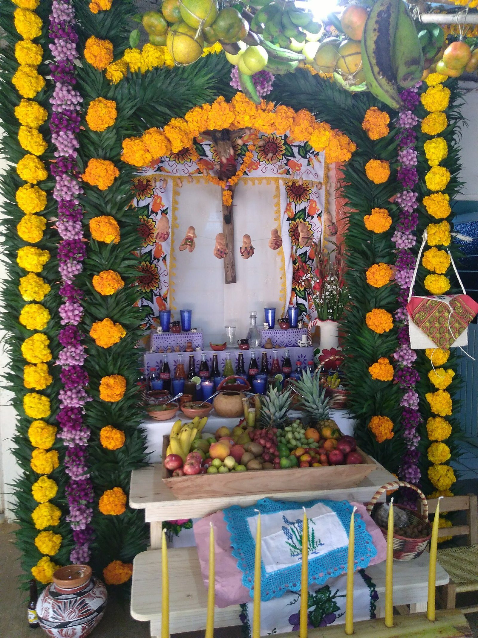 Altar de muertos con arcos de cempasúchil y flores moradas, frutas, velas y una cruz al centro