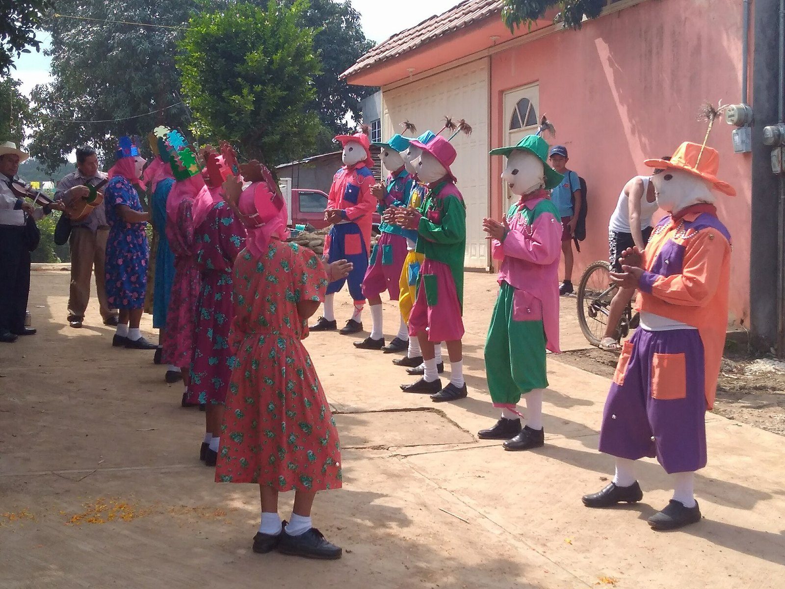 Grupo de danzantes con máscaras blancas y trajes rosas, verdes y morados bailando en una calle de tierra, con músicos al fondo